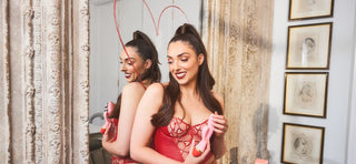 Nelly London with brown hair wearing red lingerie holding pink and red rabbit vibrator in hands and smiling against mirror with love heart drawn in red lipstick on glass