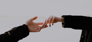 Two people's arms dressed in black jackets reaching outstretched hands to gently touch each other's fingertips