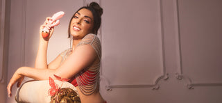Nelly London with brown hair wearing red bra and gold chain bolero resting on armchair holding pink and red rabbit vibrator in right hand while smiling
