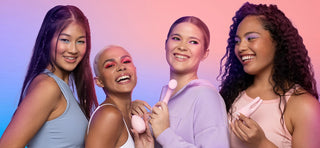 Four young women standing and smiling against gradient purple/blue/pink background. Three women hold pink vibrators from VUSH Pop Collection.