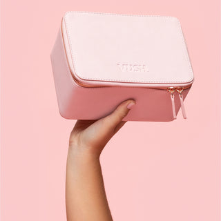 Hand holding up pink hard storage case with closed rose gold zip and text that states "VUSH" against light pink background
