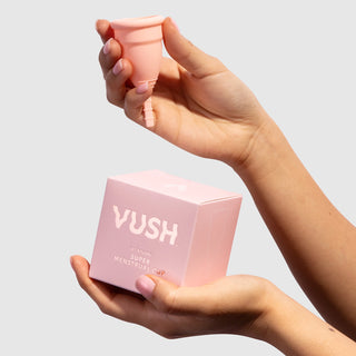 Left hand holding pink square box that reads "VUSH LET'S FLOW SUPER MENSTRUAL CUP" below right hand holding pink menstrual cup against light grey background