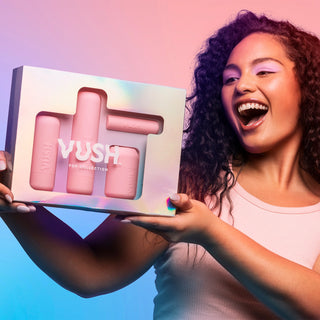 Young woman with long dark curly hair smiling widely and looking at iridescent box packaging labelled "VUSH POP COLLECTION" with clear window to display four vibrator cases inside