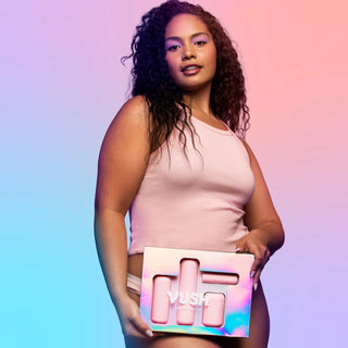 Young woman with long dark curly hair looking directly into camera and holding iridescent box packaging labelled "VUSH POP COLLECTION" with clear window to display four vibrator cases inside